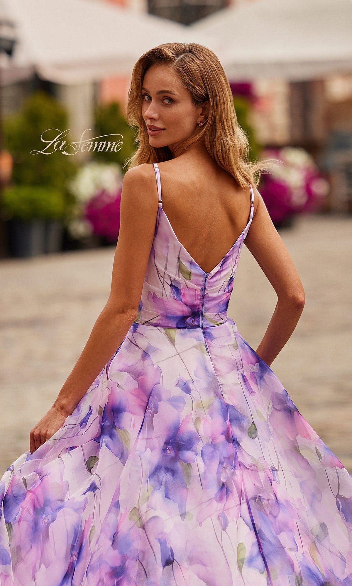 Woman wearing the La Femme 33606 long prom dress, featuring a floral print chiffon fabric, adjustable spaghetti straps, and an a-line skirt, stands outdoors on a stone path and looks over her shoulder.